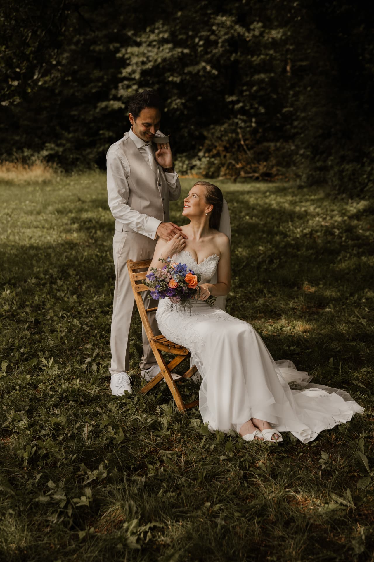 Bruid op een houten stoel in het gras met bruidegom achter haar, trouwfotografie Achterhoek