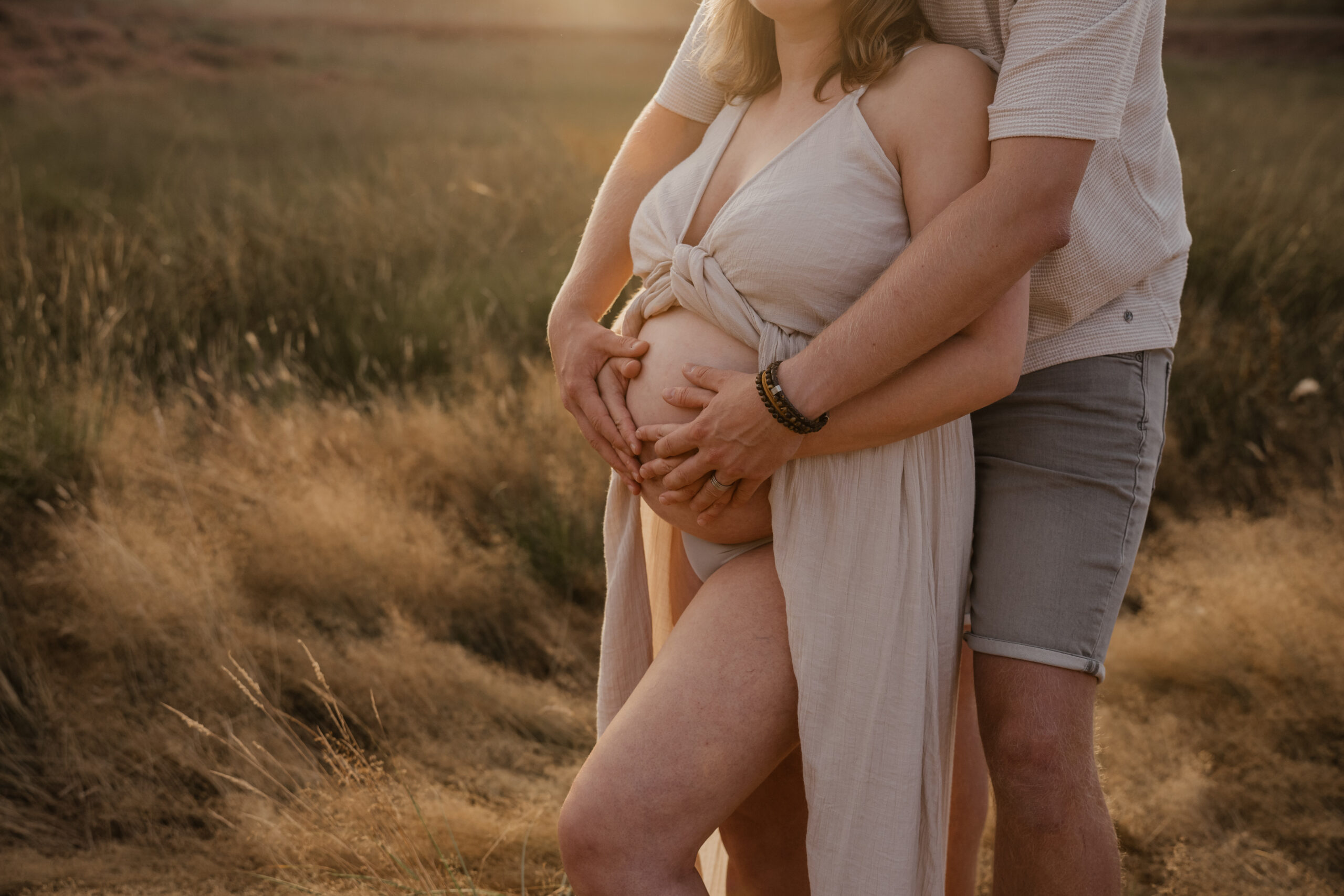 Handen van beide ouders om de zwangere buik in het hoge gras